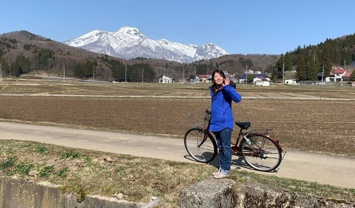 飯田さんの奥さんが自転車から降りて手を振っている。その奥には雪山が見える。